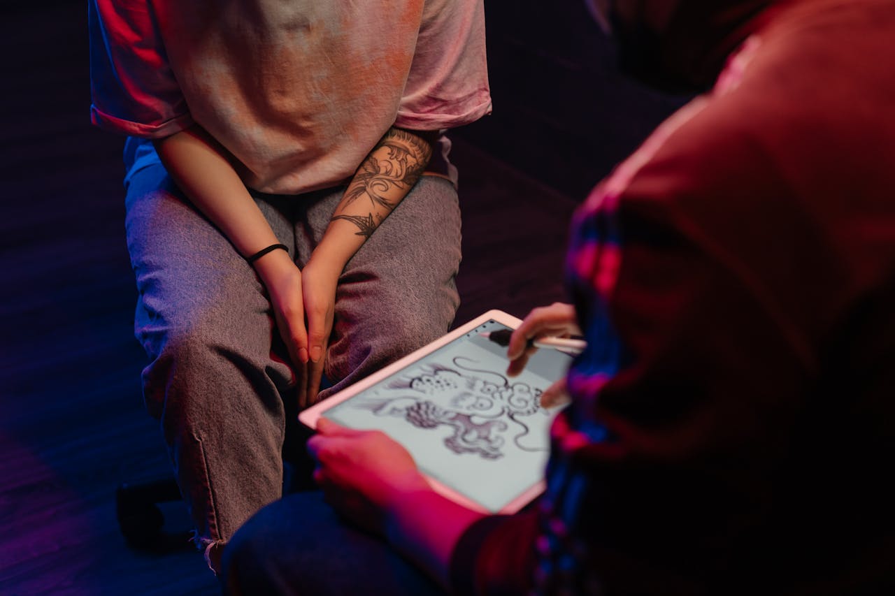 A tattoo artist and client discussing a dragon design on a digital tablet in a dimly lit tattoo studio.